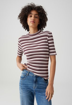 Frau mit lockigem Haar, die ein kurzärmeliges, bordeaux-weiß gestreiftes Oberteil und blaue Jeans trägt, steht vor einem schlichten hellen Hintergrund.