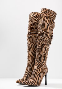 Botas de tela hasta la rodilla con frunces en color marrón con rayas negras de cebra. La punta afilada y el delgado tacón de aguja aportan altura y elegancia.