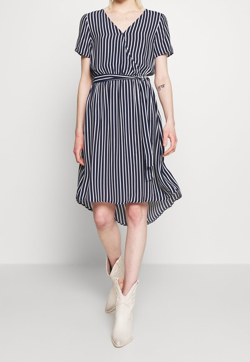 Navy and white striped dress with short sleeves, wrap design, and a high-low hem. Model wearing beige cowboy boots.