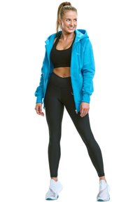 Tirkizna zip-up dukserica, crni sportski grudnjak i tajice visokog struka. Ima opušteni kroj i flis teksturu. Uparen s bijelim sportskim cipelama.
