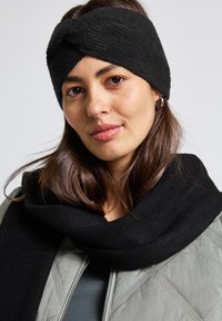 Diadema de punto negra con un diseño retorcido, combinada con una bufanda negra suelta, llevada sobre una chaqueta verde acolchada. Textura suave y material cálido.
