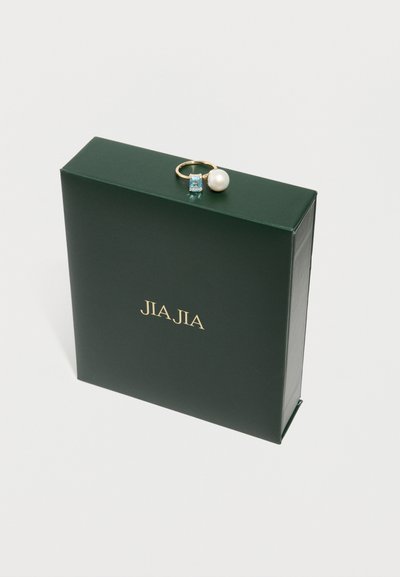 Bague en or avec une pierre précieuse bleue rectangulaire et une perle reposant sur une boîte vert foncé avec le texte doré "JIA JIA" sur une surface blanche.
