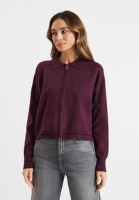 Bordeaux kleurige ritssluiting cardigan met een kraag, geribbelde rand en manchetten, gemaakt van zachte stof. Gecombineerd met lichtgrijze jeans.