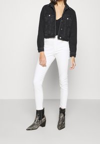 Chaqueta recortada de mezclilla negra, jeans ajustados blancos y botas de tobillo con un patrón negro y de serpiente. Líneas limpias y silueta ajustada.