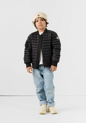 Giubbotto a raglan nero con chiusura lampo e polsini a costine, indossato sopra una camicia bianca, abbinato a jeans azzurri chiari e sneakers beige.