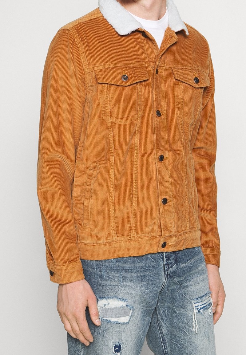 Veste en velours côtelé orange-brun avec un col en fausse fourrure blanche. Comprend des poches avant et des boutons noirs. Portée avec un jean bleu déchiré.