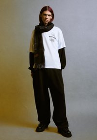 T-shirt bianca con stampa grafica, sovrapposta a una maglietta a maniche lunghe nera. Pantaloni neri a gamba larga e sciarpa a quadri. Scarpe nere.