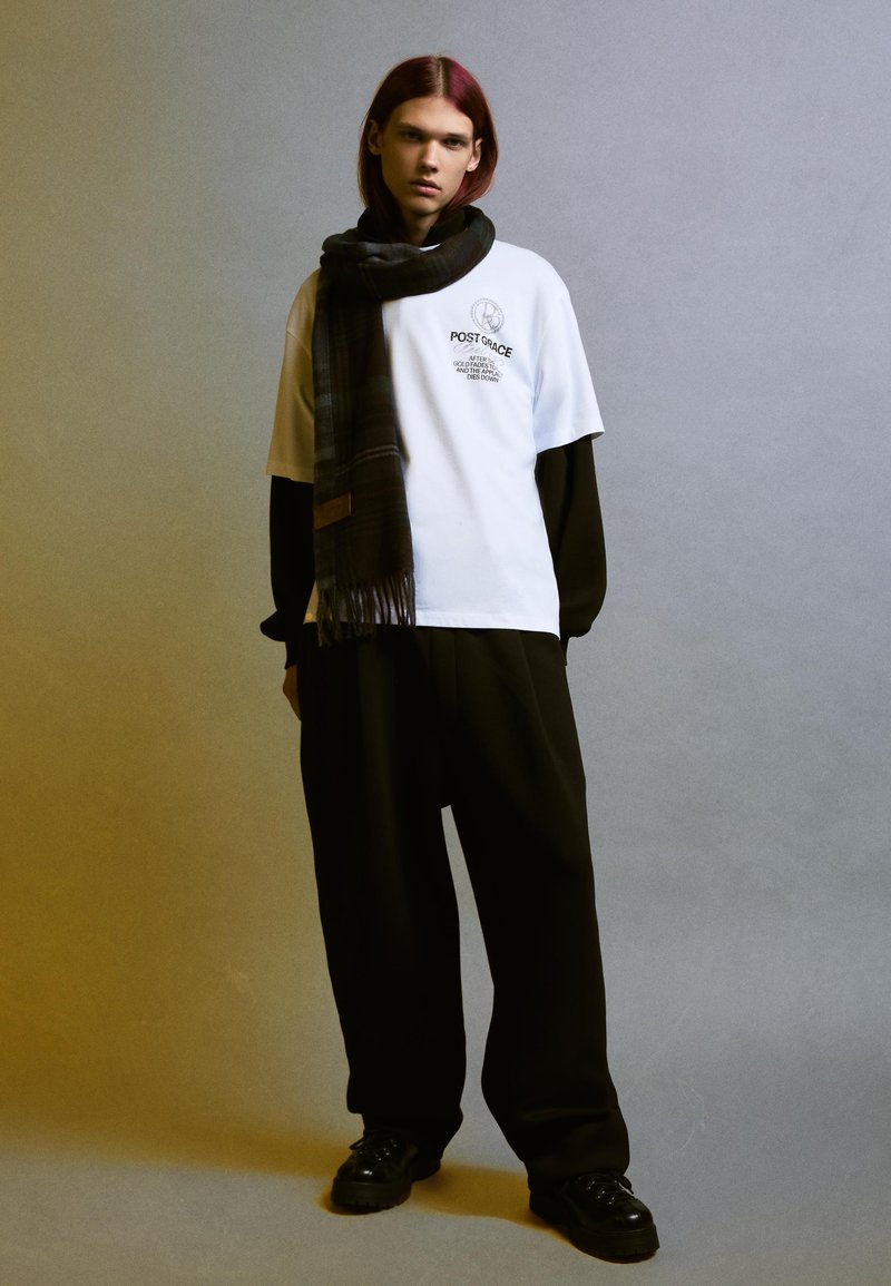 T-shirt bianca con stampa grafica, sovrapposta a una maglietta a maniche lunghe nera. Pantaloni neri a gamba larga e sciarpa a quadri. Scarpe nere.