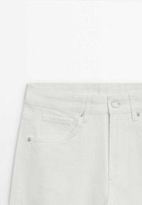 Partie supérieure d’un jean en denim blanc montrant la ceinture, la fermeture par bouton, les passants de ceinture et la poche avant avec rivets sur fond blanc.
