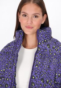 Fialová podšitá bunda s leopardím vzorem, s černými akcenty a zelenými skvrnami, vysoký límec a zapínání na zip.