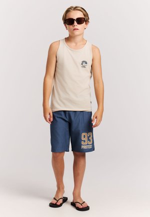 Débardeur beige avec un logo bleu, associé à un short en mesh bleu foncé avec un "93" doré et le texte "PROTEST". Tongs noires et lunettes de soleil.
