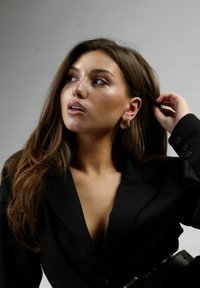 Blazer negro con cuello abierto, ceñido a la cintura con un cinturón. Grandes pendientes de aro dorados. Cabello largo y liso de color castaño. Fondo neutro.