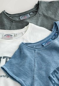 Trois sweatshirts Superdry pliés en gris, blanc et bleu, montrant les encolures et les étiquettes de la marque.