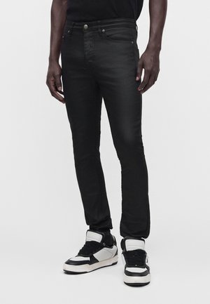 Homme portant un jean noir slim et des baskets noir et blanc, debout devant un fond uni.