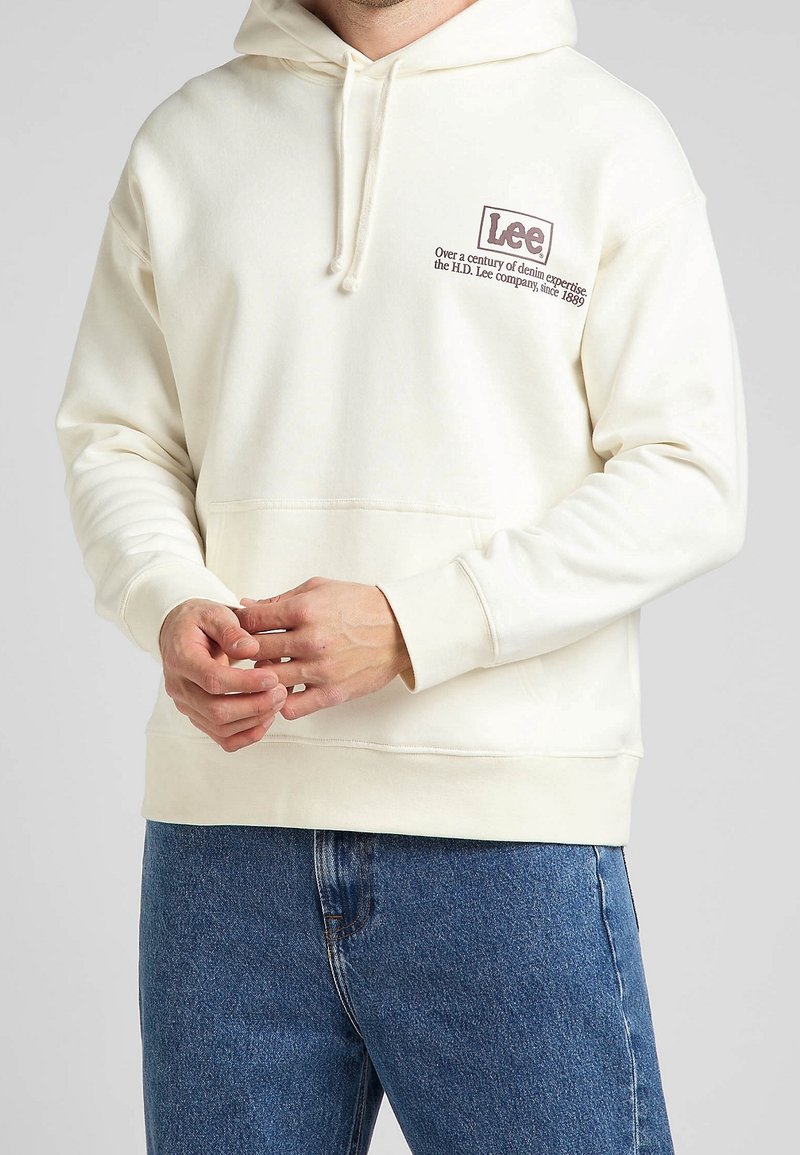 Sudadera crema con un bolsillo canguro y puños acanalados. Presenta un logotipo impreso en marrón, mostrando un diseño casual y de corte holgado.