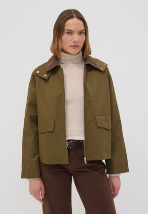 Frau trägt olivgrüne Jacke mit braunem Kragen, beigen Rollkragenpullover, braune Hose und schwarzen Gürtel, steht vor einem einfarbigen Hintergrund.