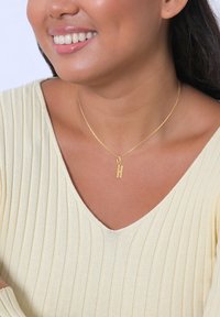Collier en or mettant en valeur un pendentif texturé en forme de lettre "H", porté par une personne vêtue d'un haut crème à côtes légères.