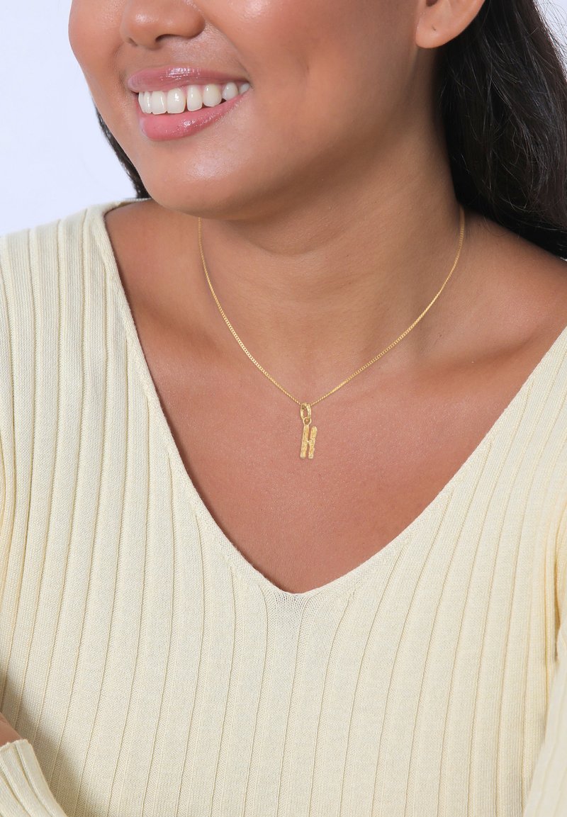 Collier en or mettant en valeur un pendentif texturé en forme de lettre "H", porté par une personne vêtue d'un haut crème à côtes légères.