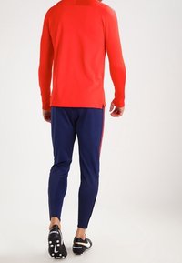 Chemise de sport rouge à manches longues, associée à un pantalon ajusté bleu avec des bandes latérales rouges. Chaussures de sport noires avec plusieurs crampons sous la semelle.