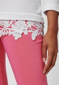 Roze fitted broek met witte kanten bloemenrand op de taille. De stof is glad; de kant heeft ingewikkelde bloemmotieven en details.