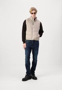 Beige quilted vest with a high collar, paired with a dark brown long-sleeve shirt, blue jeans, and black shoes. Sunglasses complete the look.