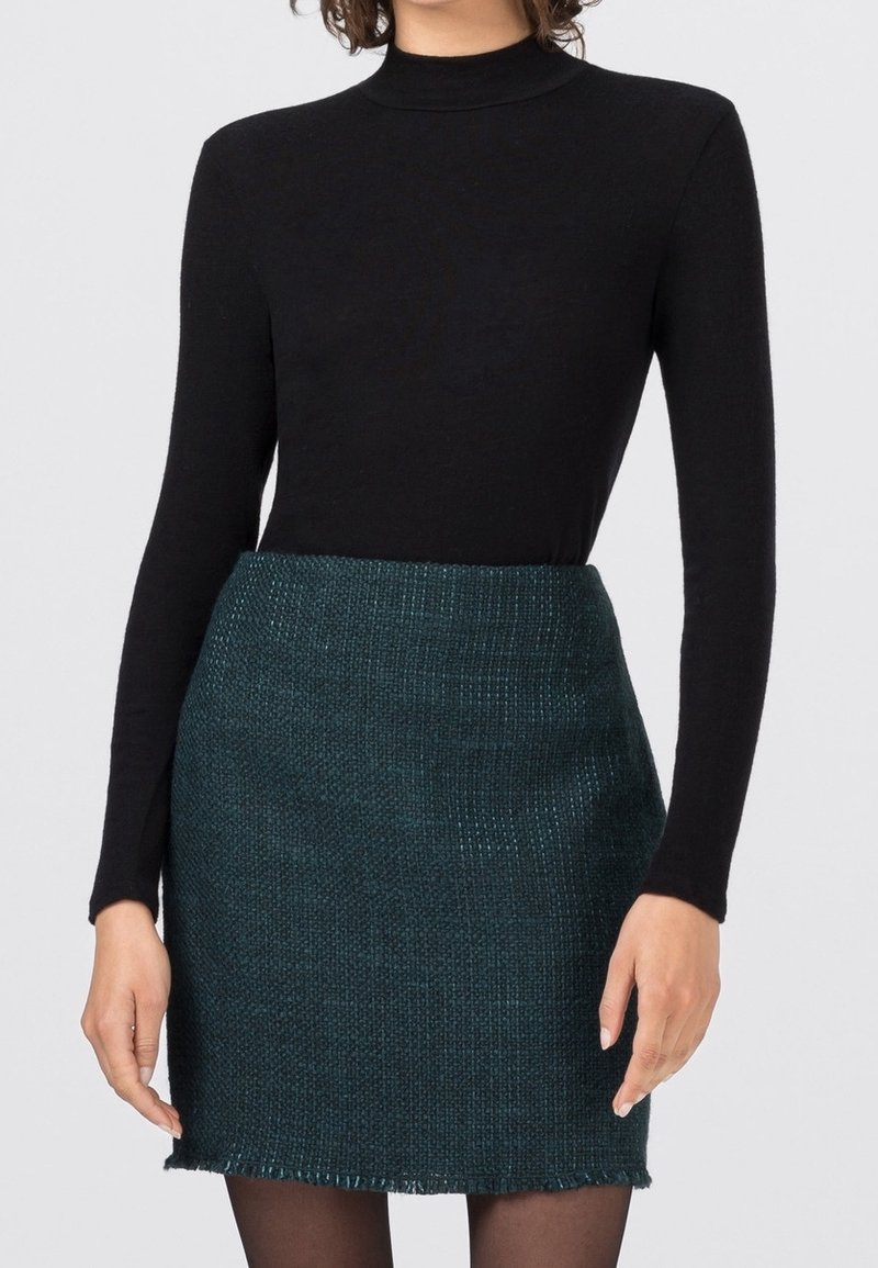 Femme portant un haut noir à manches longues col montant et une mini-jupe vert foncé texturée avec des collants noirs, debout devant un fond clair.