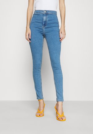 Kvinde set fra taljen og ned iført blå skinny jeans med høj talje og gule sandaler med hæl, stående mod en ensartet lys baggrund.