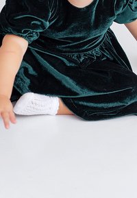Zelené sametové šaty s vypuštěnými rukávy, nabíraným pasem a bílými ponožkami s krajkovým vzorem. Zaměření na dolní část těla, sedící na bílé ploše.