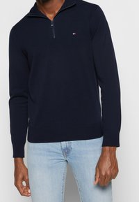 Pull zippé bleu marine en tissu doux, doté d'un col montant et d'un petit logo sur la poitrine. Manches longues et ourlet côtelé.