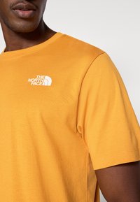 Κίτρινο βαμβακερό t-shirt με κοντά μανίκια, με λευκό λογότυπο στο αριστερό στήθος. Το ύφασμα φαίνεται μαλακό με λεία υφή.