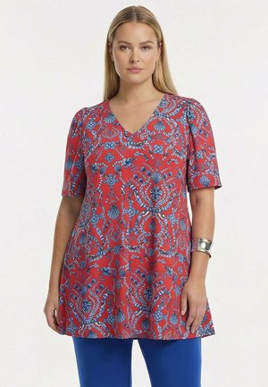 Kvinde med glat blondt hår iført en rød tunika med blå blomster- og skalmønstre samt blå bukser, stående foran en ensfarvet hvid baggrund.