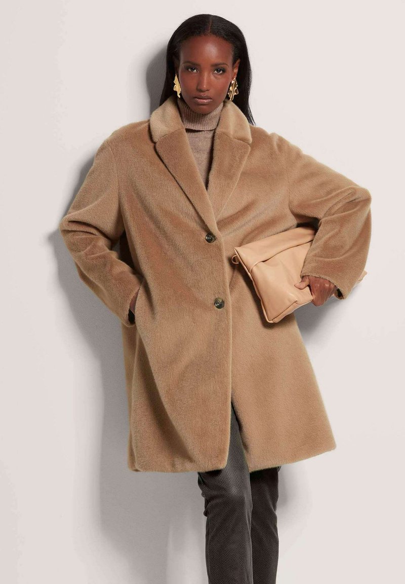 Camelkleurige wollen jas met een sjawlkragen, twee knopen en zijzakken. Model houdt een beige leren clutch vast. Zachte textuur en relaxte fit.