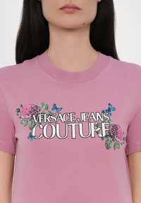 Rosa bomull T-skjorte med blomster- og sommerfuglgrafikk, og teksten "VERSACE JEANS COUTURE" i fet svart skrift på forsiden.