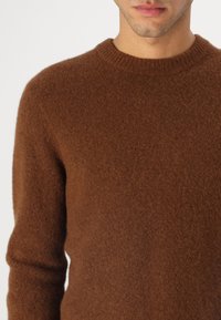 Pulover din lână maro, cu un gât rotund, finisaj texturat și mâneci lungi. Țesătura pare moale și călduroasă, punând accent pe un design confortabil.