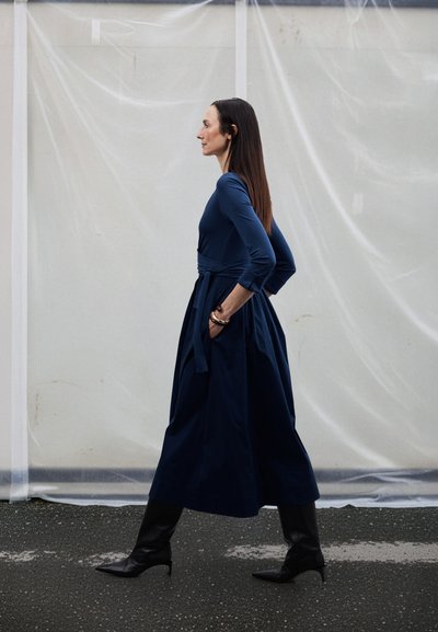 Robe longue bleu marine avec un corsage ajusté et une jupe large et fluide. La robe est dotée d'une taille nouée et est associée à des bottes noires montant jusqu'aux genoux.