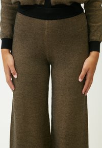 Bruine, textuurrijke wijde broek met een subtiel visgraatpatroon. Elastische tailleband met zwarte ribbel. Handen op de heupen geplaatst.