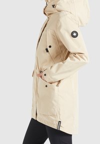 Beżowa wodoodporna parka z kapturem, bocznymi kieszeniami oraz czarną naszywką z logo na lewym rękawie; gładka tkanina o lekko błyszczącej fakturze.