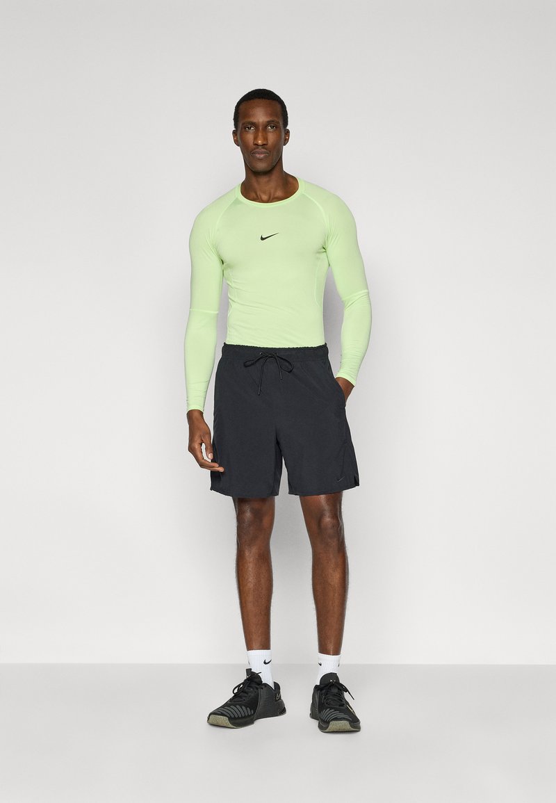 Hellgrünes langärmeliges Sportoberteil mit schwarzem Nike-Logo, kombiniert mit schwarzen Shorts und schwarzen Sportschuhen mit grauen Akzenten.