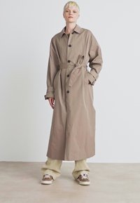Beige trench kabát, derekán megkötővel, gallérja és hosszú ujjai vannak, világos árnyalatú, bőszárú nadrággal és sötét tornacipővel párosítva.