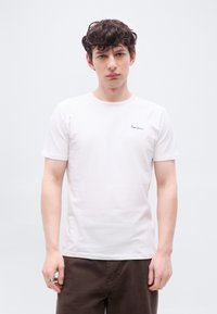 Νεαρός με καστανά σγουρά μαλλιά, που φοράει μια απλή λευκή κοντομάνικη μπλούζα και σκούρο καφέ παντελόνι, μπροστά σε λευκό φόντο.