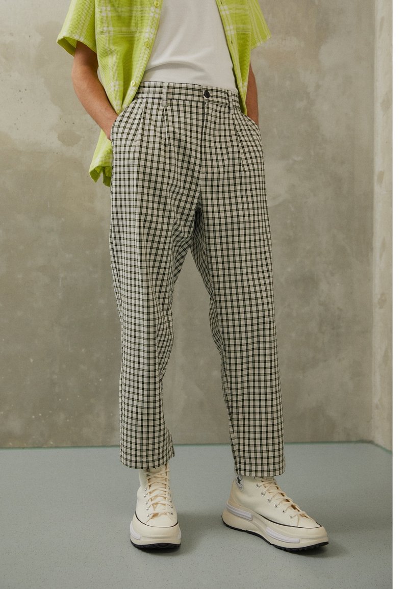 Person wearing checkered trousers, white high-top sneakers, white shirt, and green short-sleeve button-up shirt, standing against gray wall.