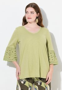 Maglietta verde chiaro a V con maniche tre quarti, caratterizzata da dettagli in occhio di pernice sui polsini. Tessuto morbido e vestibilità comoda, abbinata a pantaloni stampati.