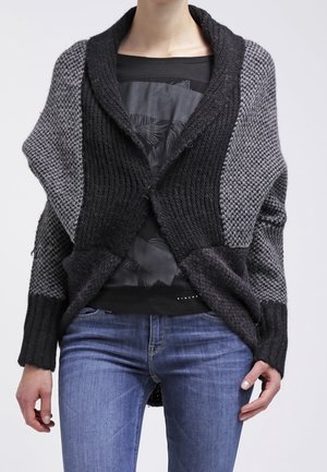 Personne portant un cardigan en grosse maille avec des blocs de couleurs noir et gris par-dessus une chemise noire et un jean bleu.