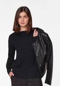 Pull en maille noire à motif texturé, associé à une veste en cuir noire avec des fermetures à pression. Le mannequin pose avec une main sur la hanche.