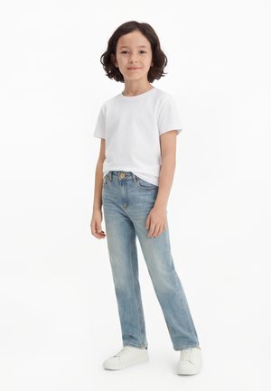 Barn med middels langt mørkt hår iført hvit t-skjorte, lys blå jeans og hvite joggesko, står mot hvit bakgrunn.