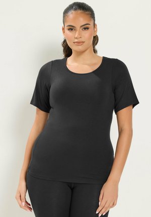 Femme aux longs cheveux foncés attachés, portant un haut noir ajusté à manches courtes et un pantalon noir, debout devant un fond clair uni.