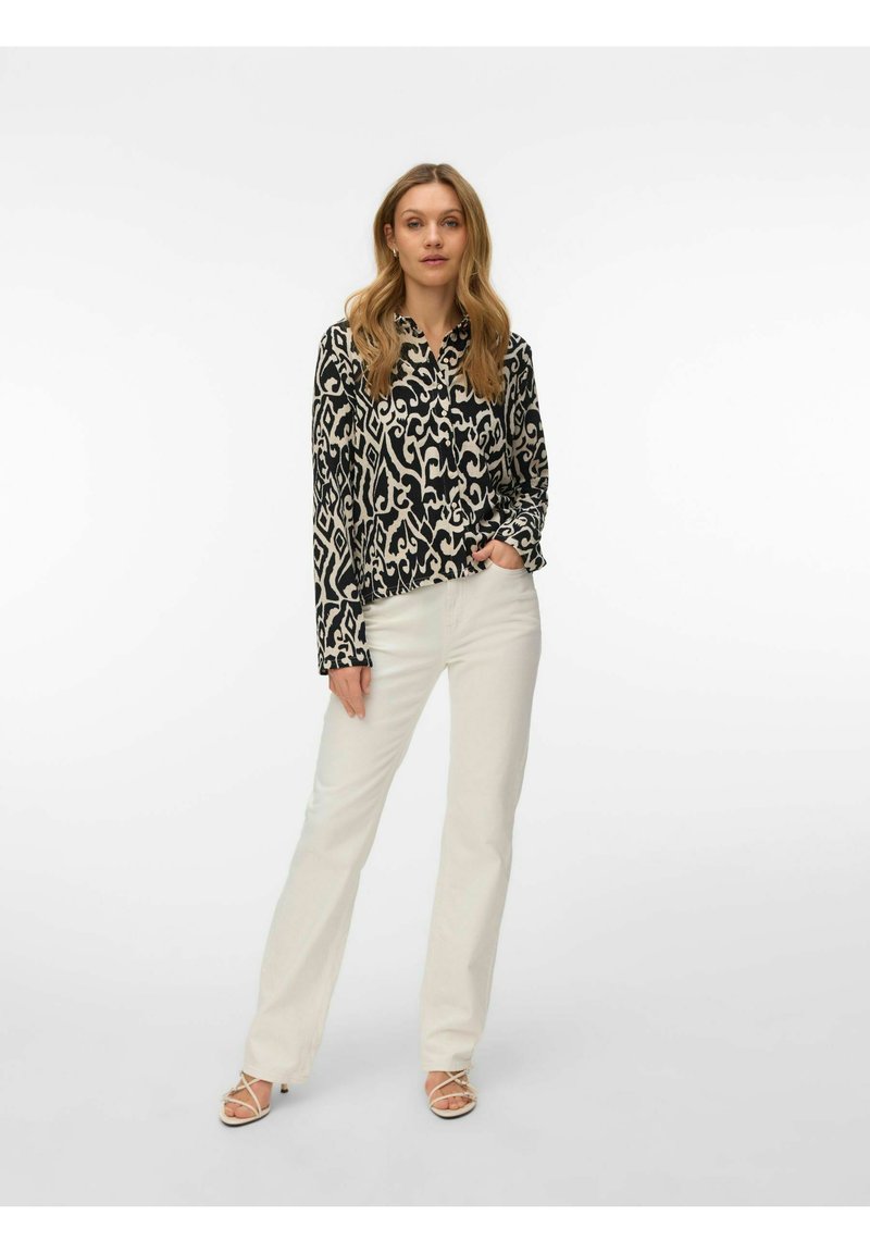 Femme debout, main dans la poche, portant une chemise à manches longues noire et blanche à motifs, un pantalon blanc et des talons blancs à brides sur un fond uni.