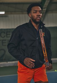 Mees mustas jope ja oranžides lühikestes pükstes hoiab siseruumides tenniseväljakul tennisesaabrit, valmistudes mängima.