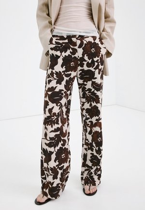 Persoon draagt wijde broek met grote donkerbruine bloemenpatronen op een lichte achtergrond, gecombineerd met een beige blazer en zwarte sandalen.