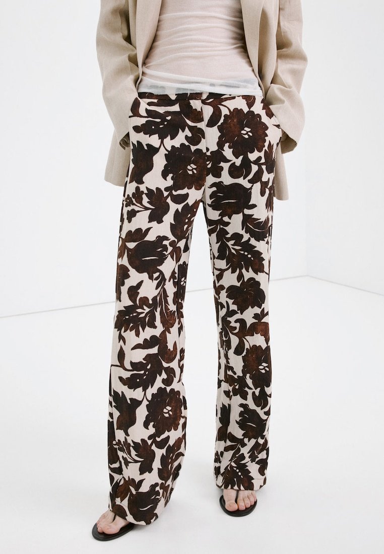 Osoba nosząca szerokie spodnie z dużym, ciemnobrązowym kwiatowym wzorem na jasnym tle, w połączeniu z beżową marynarką i czarnymi sandałami.